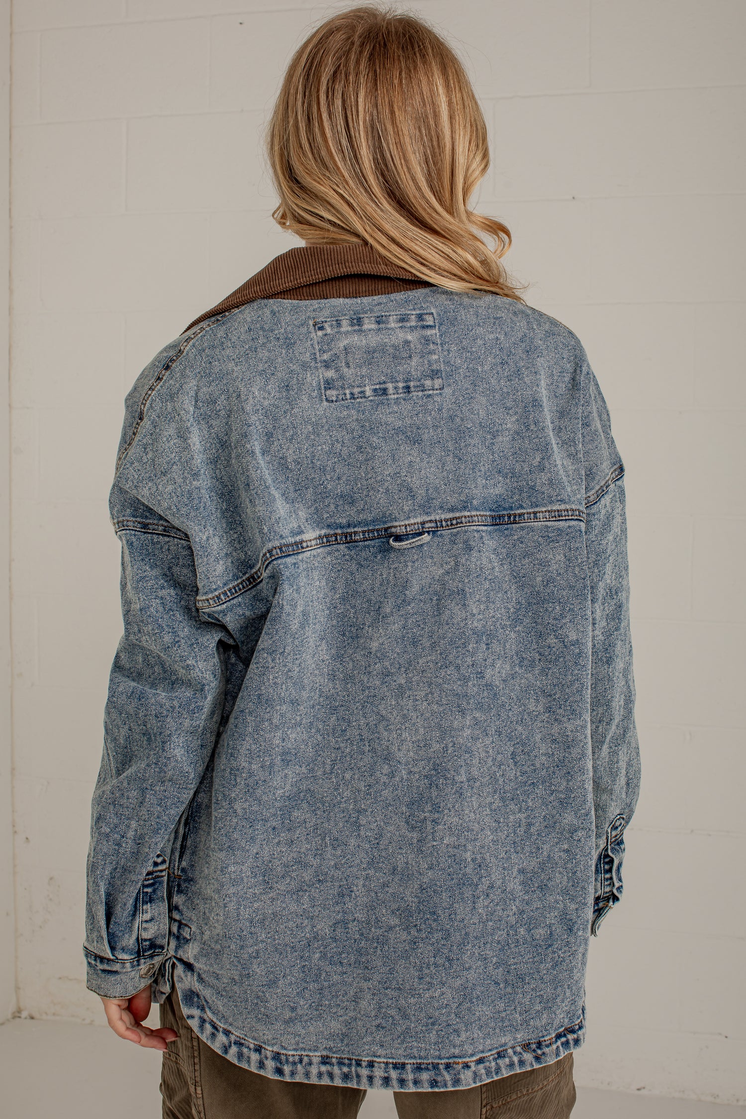 A person viewed from behind wearing an oversized washed blue denim jacket with a brown corduroy collar, paired with olive pants, against a light off-white brick wall.