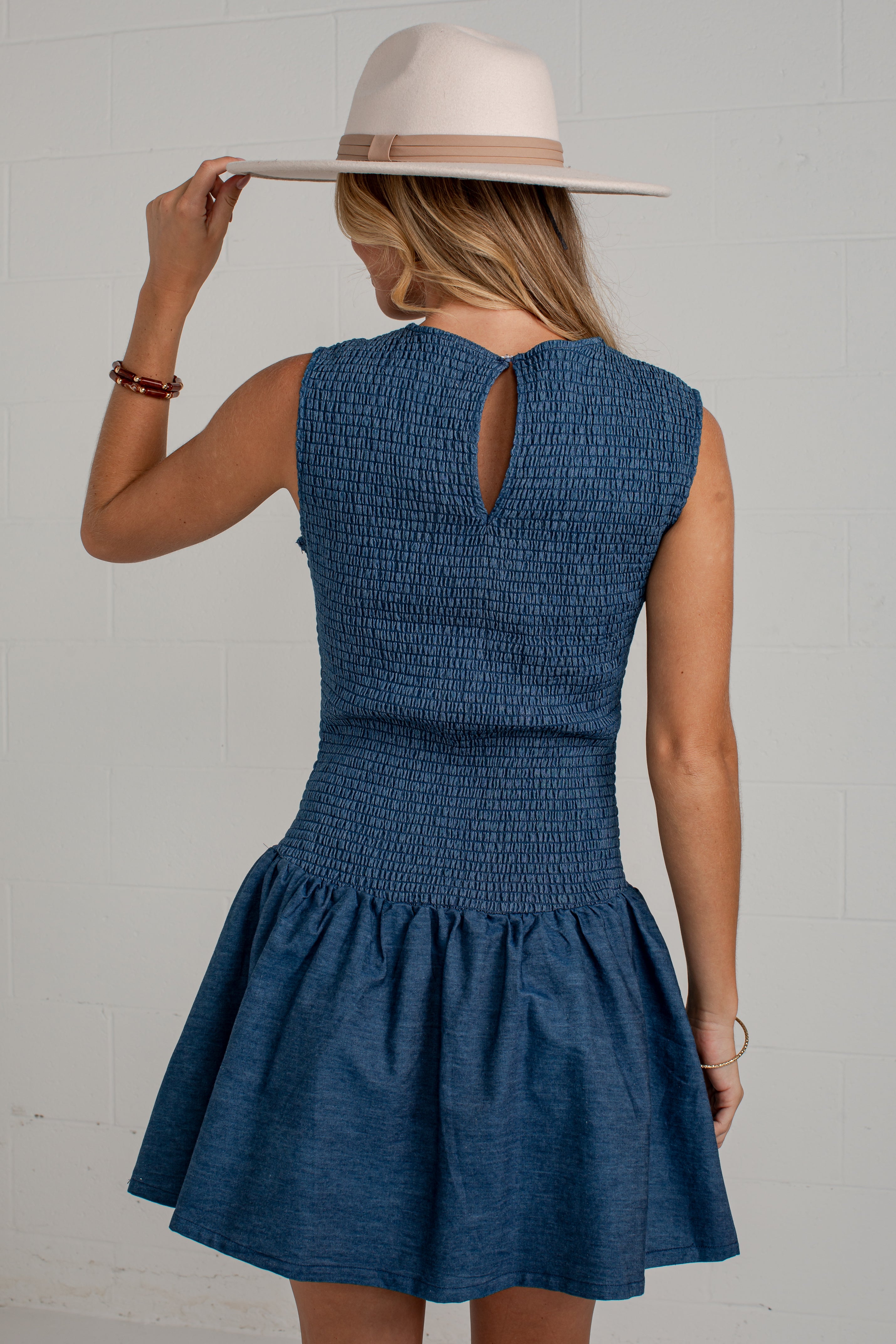A woman wearing a sleeveless blue smocked dress with a flared skirt, shown from the back while wearing a wide-brimmed cream hat against a white brick wall.