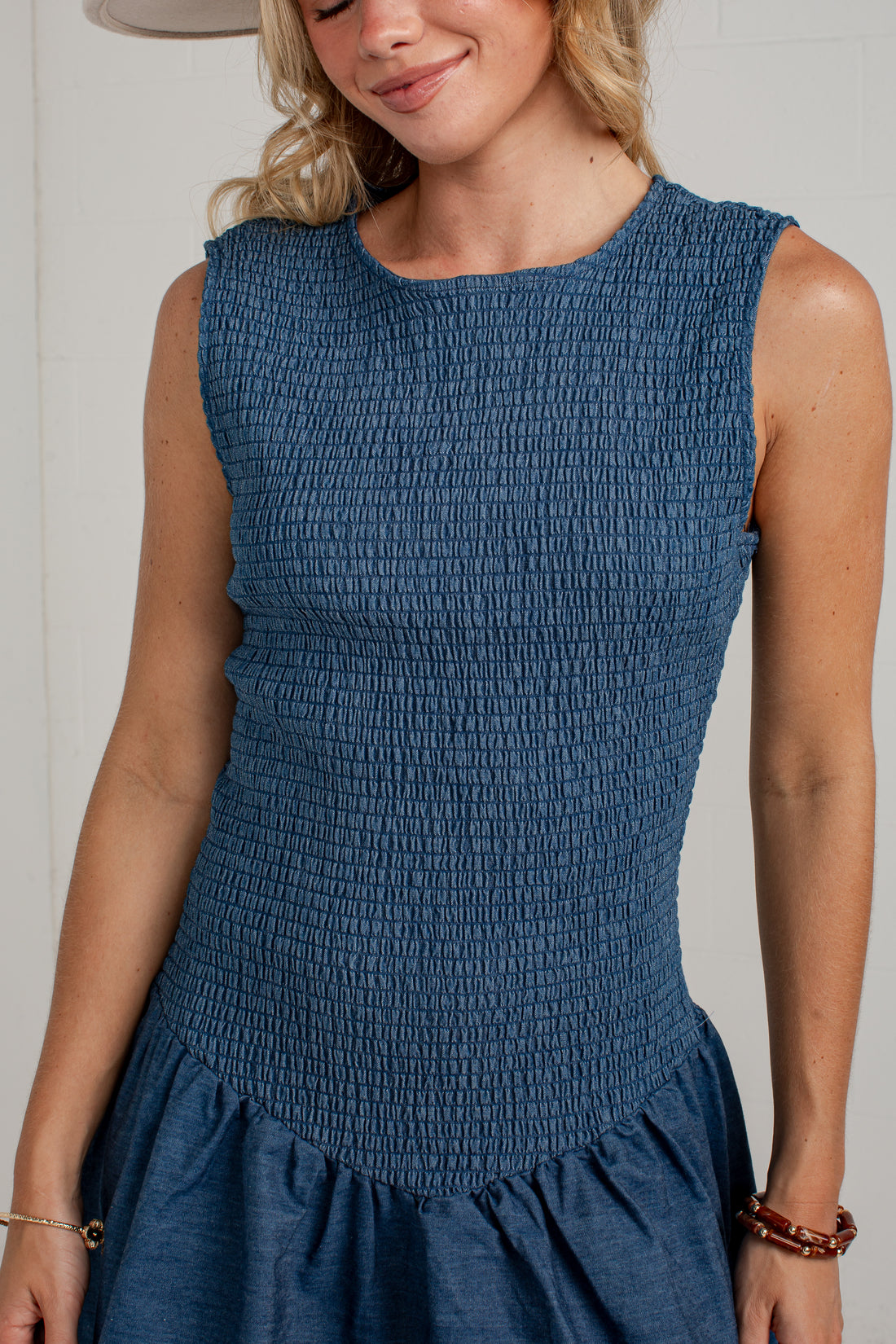 A woman wearing a sleeveless smocked blue dress, cropped from the chin down, against a plain light background.