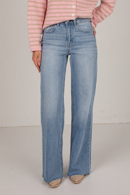 A person wearing light-wash blue wide-leg jeans, a pink and white striped long-sleeve top, and beige pointed-toe flats, standing against a plain off-white studio background.