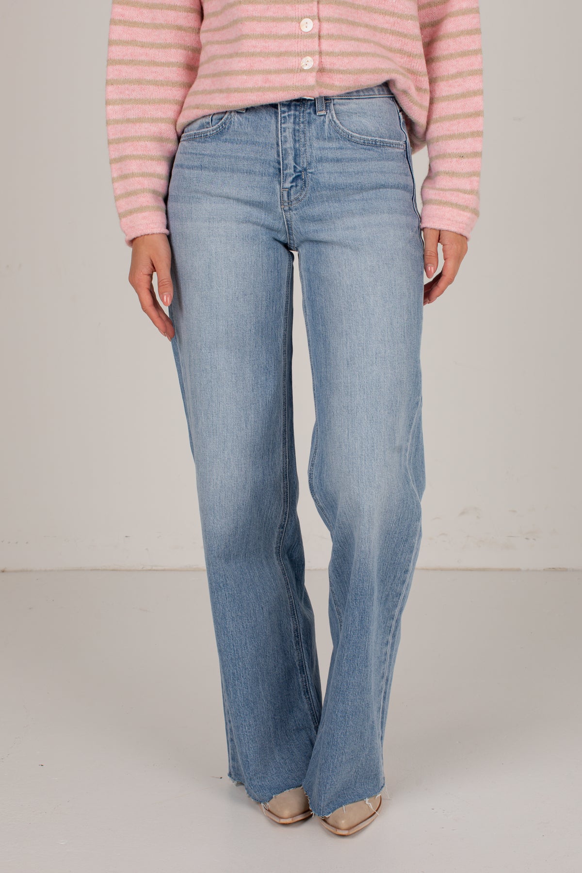 A person wearing light-wash blue flare jeans, a pink and white striped cardigan, and beige pointed-toe flats, standing against a plain off-white studio background.