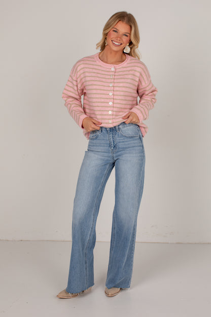 A woman modeling a pink and white striped long-sleeve button-up cardigan with light-wash blue flare jeans, standing against a plain light gray studio background.
