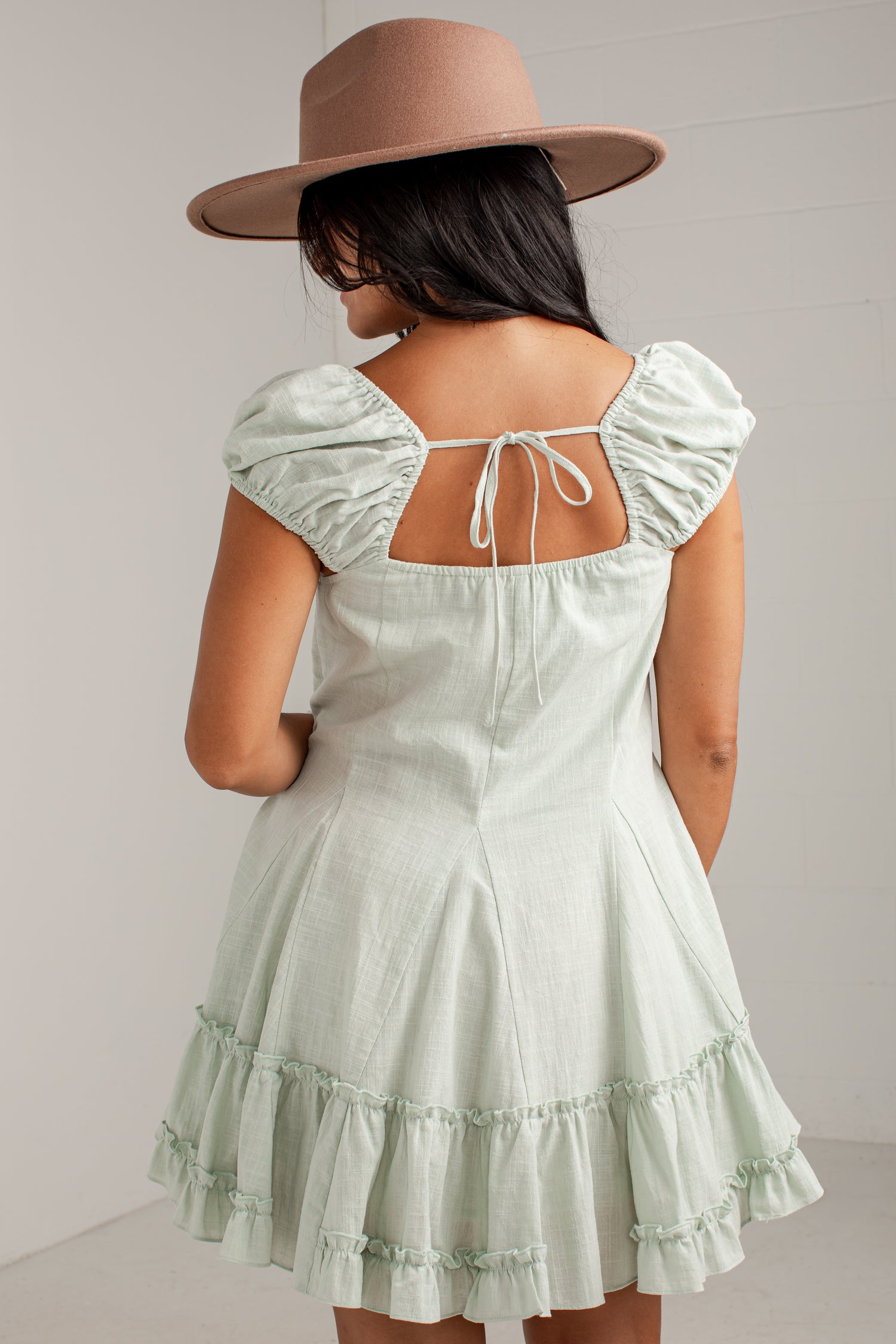 A woman shown from the back wearing a pale mint green puff-sleeve dress with a tie-back neckline and a matching wide-brim tan hat, set against a light gray wall.