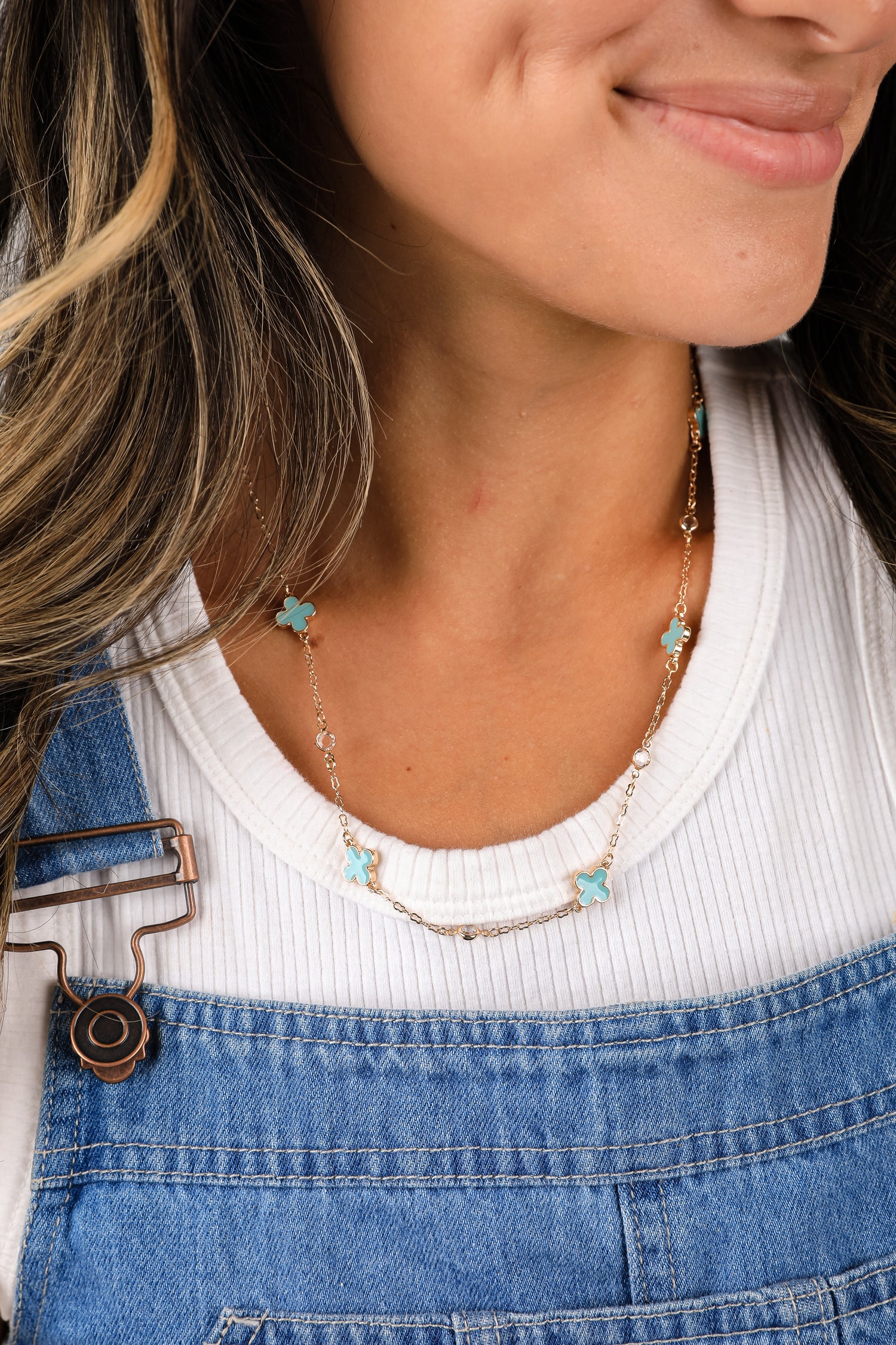 Close-up of a person wearing a necklace with turquoise heart pendants, white top, and blue denim overalls.