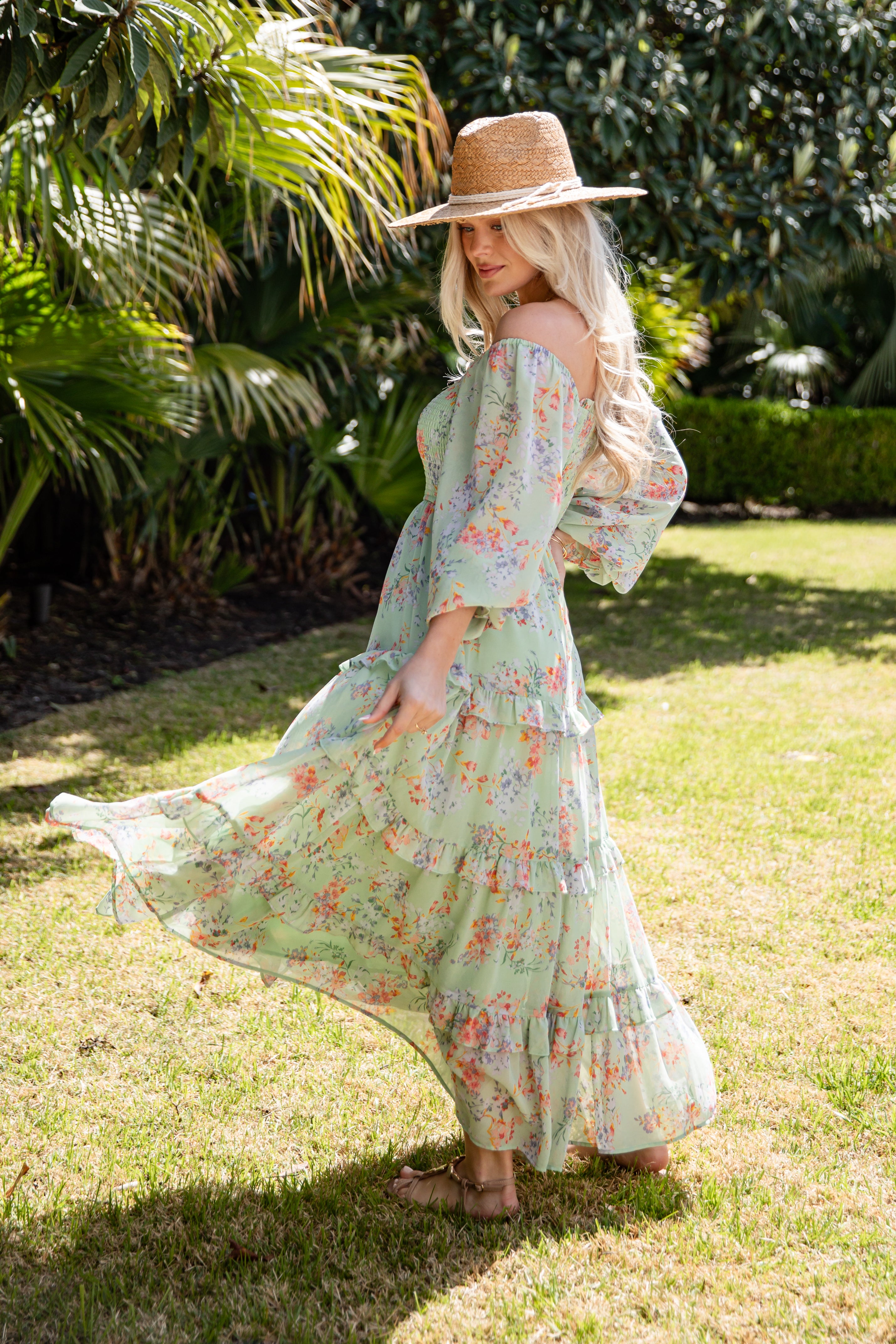 Woman in a floral dress and straw hat standing on grass with greenery in the background