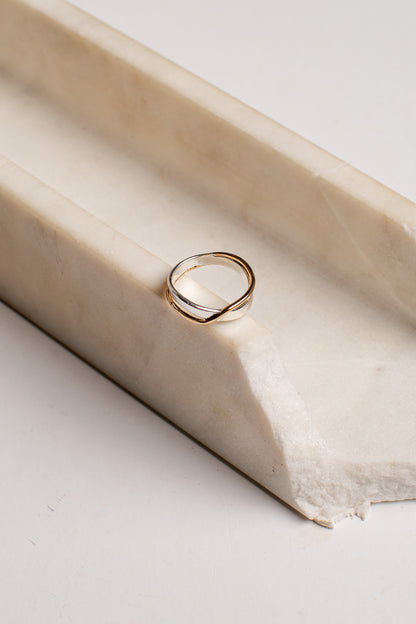 A thin double-band ring with a twisted, intertwined design in polished yellow gold, resting on a cream-colored stone riser against a light background.