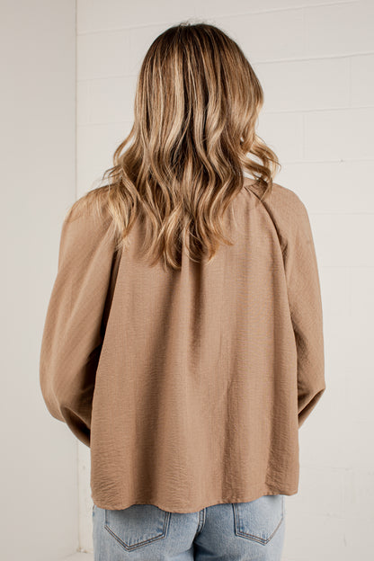A woman viewed from behind wearing a loose, long-sleeve tan top and light blue jeans, standing against a white paneled wall.