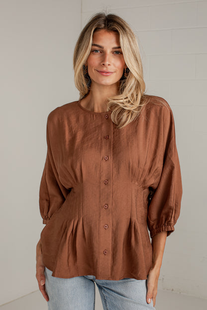 A woman wearing a rust-brown linen button-up blouse with puff sleeves and a gathered peplum waist, paired with light-wash blue jeans, standing against a light gray brick wall.