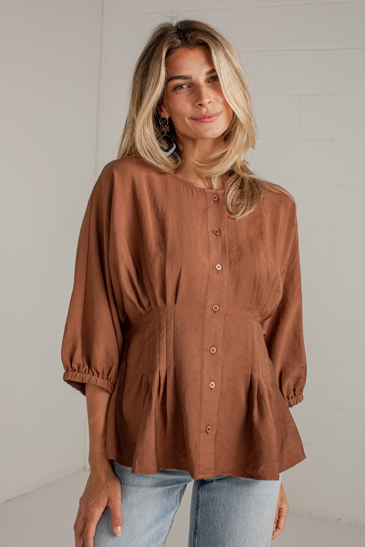 A woman wearing a rust-brown button-up blouse with three-quarter sleeves and a gathered peplum hem, paired with light-wash blue jeans, standing against a white brick wall.