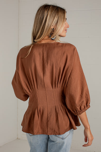 A woman viewed from the back wearing a rust-brown long-sleeve blouse with gathered puff sleeves and a peplum hem, paired with light blue jeans against a white paneled wall.