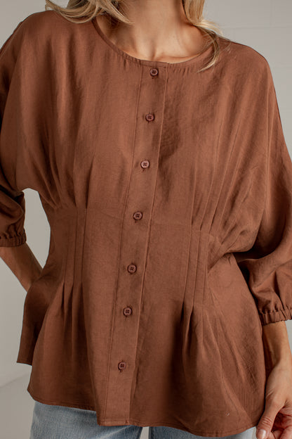 A woman wearing a brown long-sleeve button-front blouse with a round neckline and gathered peplum hem, paired with light blue jeans.