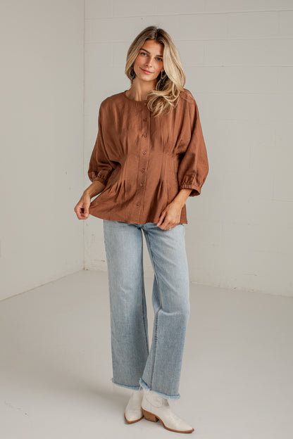 A woman modeling a brown long-sleeve button-up blouse with a gathered waist, paired with light blue jeans and white ankle boots, standing in a bright white studio.