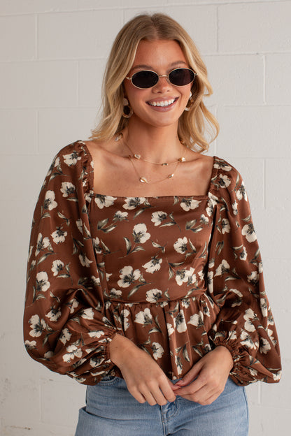 A woman wearing a brown long-sleeve blouse with a white floral print, square neckline, and puff sleeves, paired with blue jeans and round sunglasses, standing against a white brick wall.