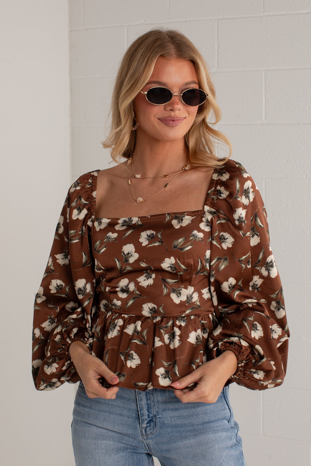 A woman wearing a brown floral puff-sleeve top with a square neckline and blue jeans, standing against a white brick wall.
