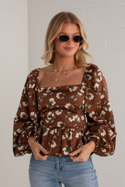A woman wearing a brown floral puff-sleeve top with a square neckline and blue jeans, standing against a white brick wall.
