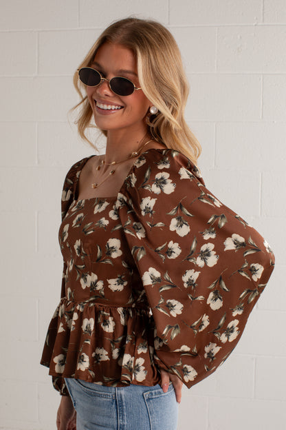 A woman wearing a brown floral puff-sleeve blouse with a peplum hem, paired with light blue jeans and oval sunglasses, standing against a white brick wall.