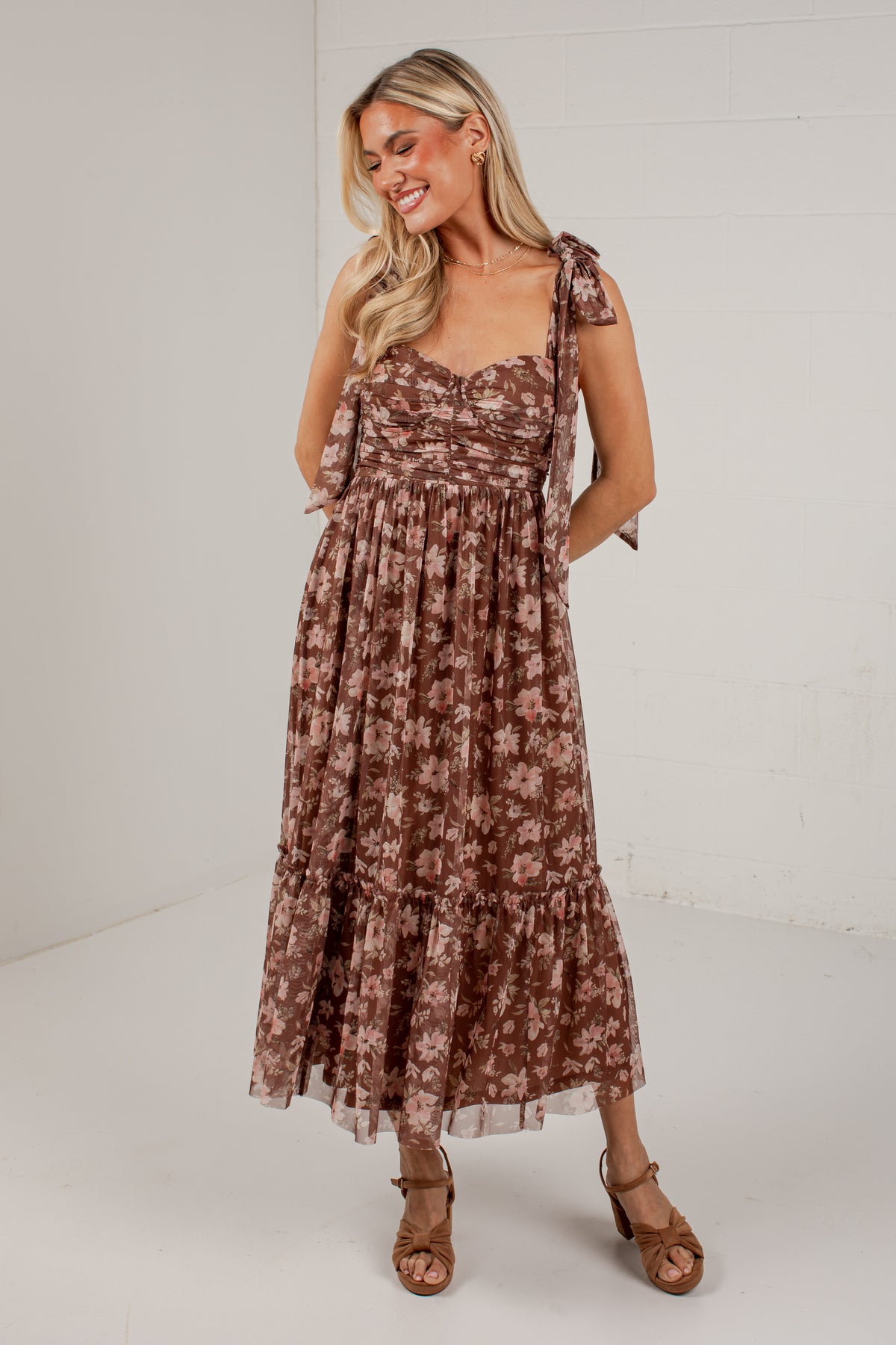 A woman modeling a brown and beige floral tiered midi dress with tie-shoulder straps and side pockets, paired with brown heeled sandals, in a bright studio setting.