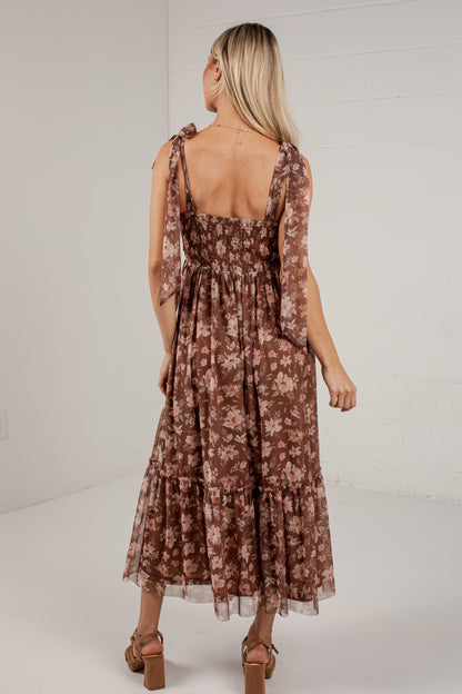 A woman wearing a brown tiered midi dress with a floral pattern, shown from the back against a white studio background.