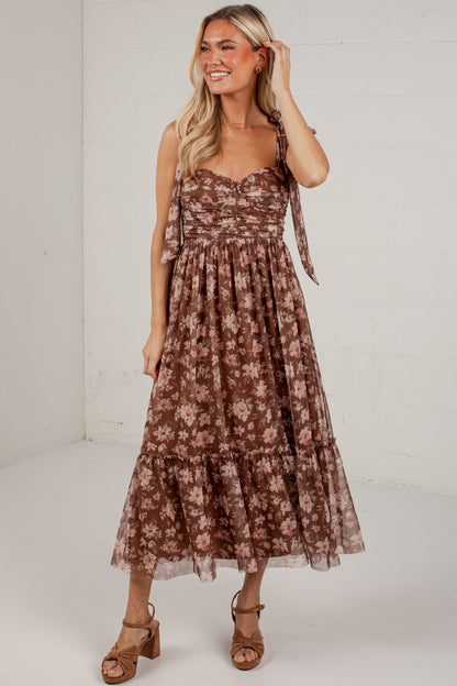 A woman modeling a brown and beige floral tiered midi dress with tie straps, paired with brown heeled sandals, in a bright white studio setting.