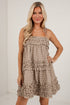 A blonde woman modeling a sleeveless tiered mini dress with a brown and beige gingham pattern, set against a white brick wall background.