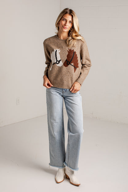 A woman modeling a taupe knit sweater with a horse graphic on the front, paired with light-wash wide-leg jeans and white ankle boots, standing in a bright studio setting.