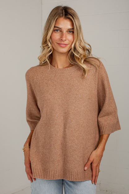 A woman wearing a soft, heathered camel-colored knit sweater with three-quarter sleeves and a crew neck, paired with light blue jeans, standing against a light gray wall.