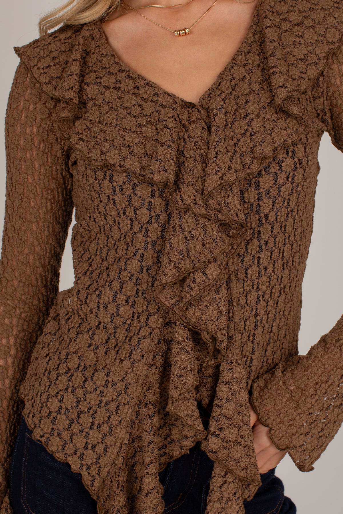 A close-up of a woman wearing a brown textured knit top with layered ruffles and long sleeves, paired with dark jeans and a delicate gold necklace.