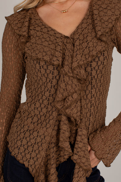 A close-up of a woman wearing a brown textured knit top with layered ruffles and long sleeves, paired with dark jeans and a delicate gold necklace.