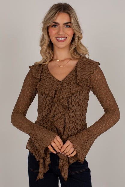 A smiling blonde woman wearing a brown textured knit long-sleeve top with ruffle details and dark blue jeans, standing against a plain light gray studio background.