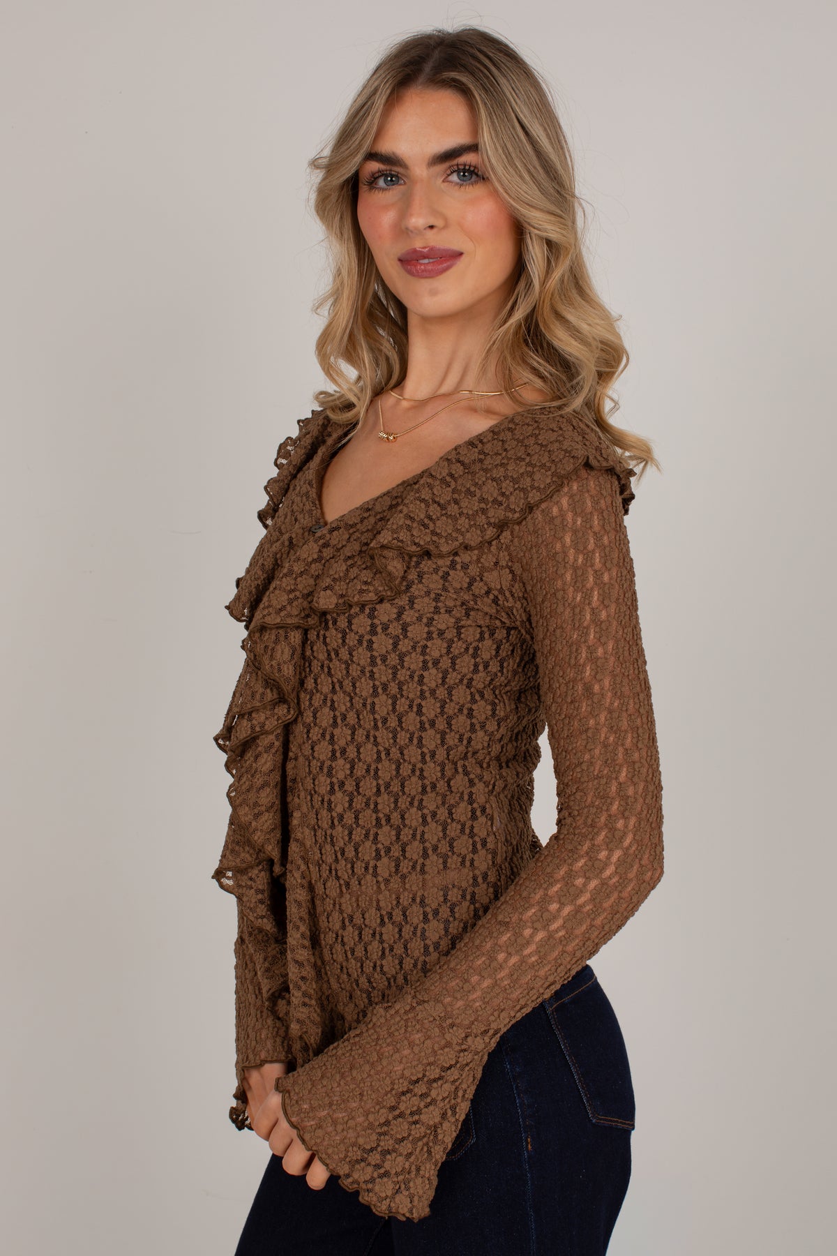 A woman wearing a brown textured knit long-sleeve top with ruffle details on the shoulders and sleeves, paired with dark blue jeans, photographed against a plain light gray background.