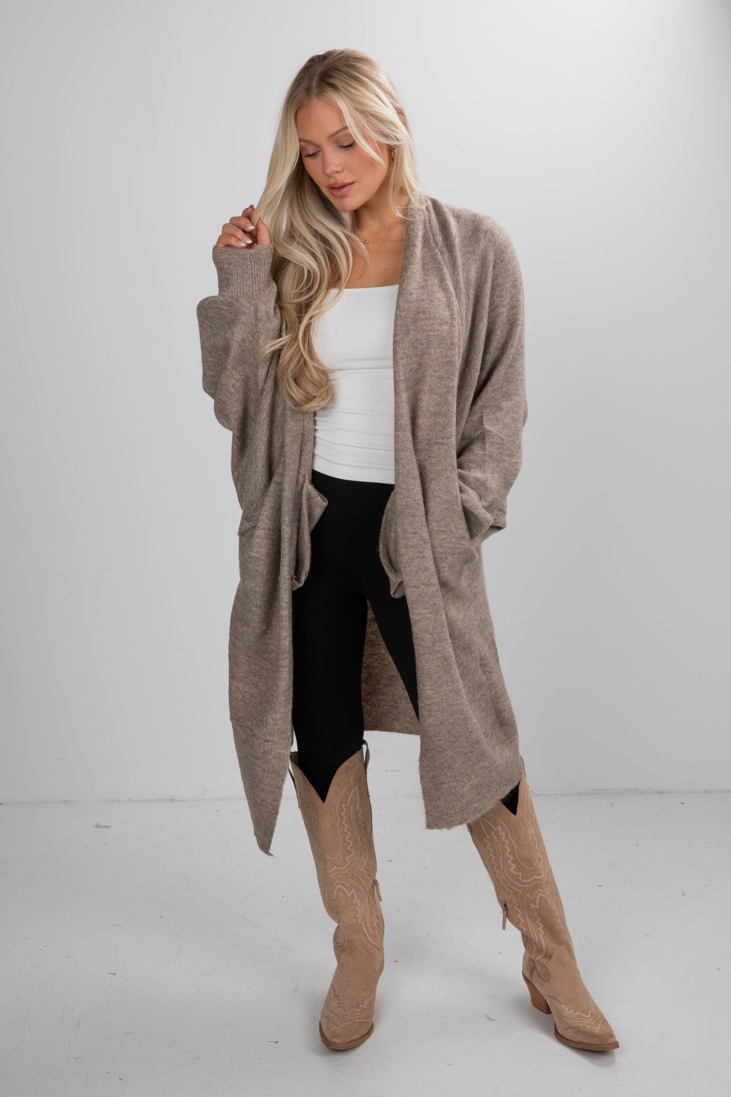 Woman wearing a long beige cardigan, white top, black leggings, and tan knee-high boots on a white background