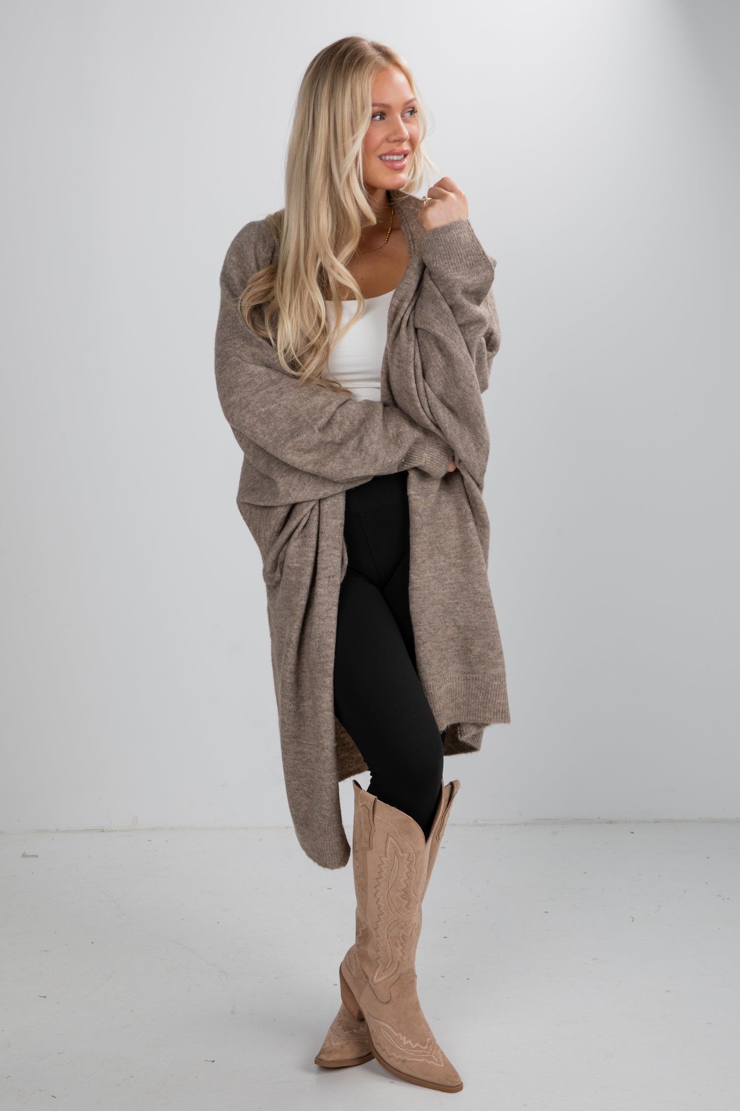 Woman wearing a long beige cardigan, white top, black pants, and tan knee-high boots on a white background