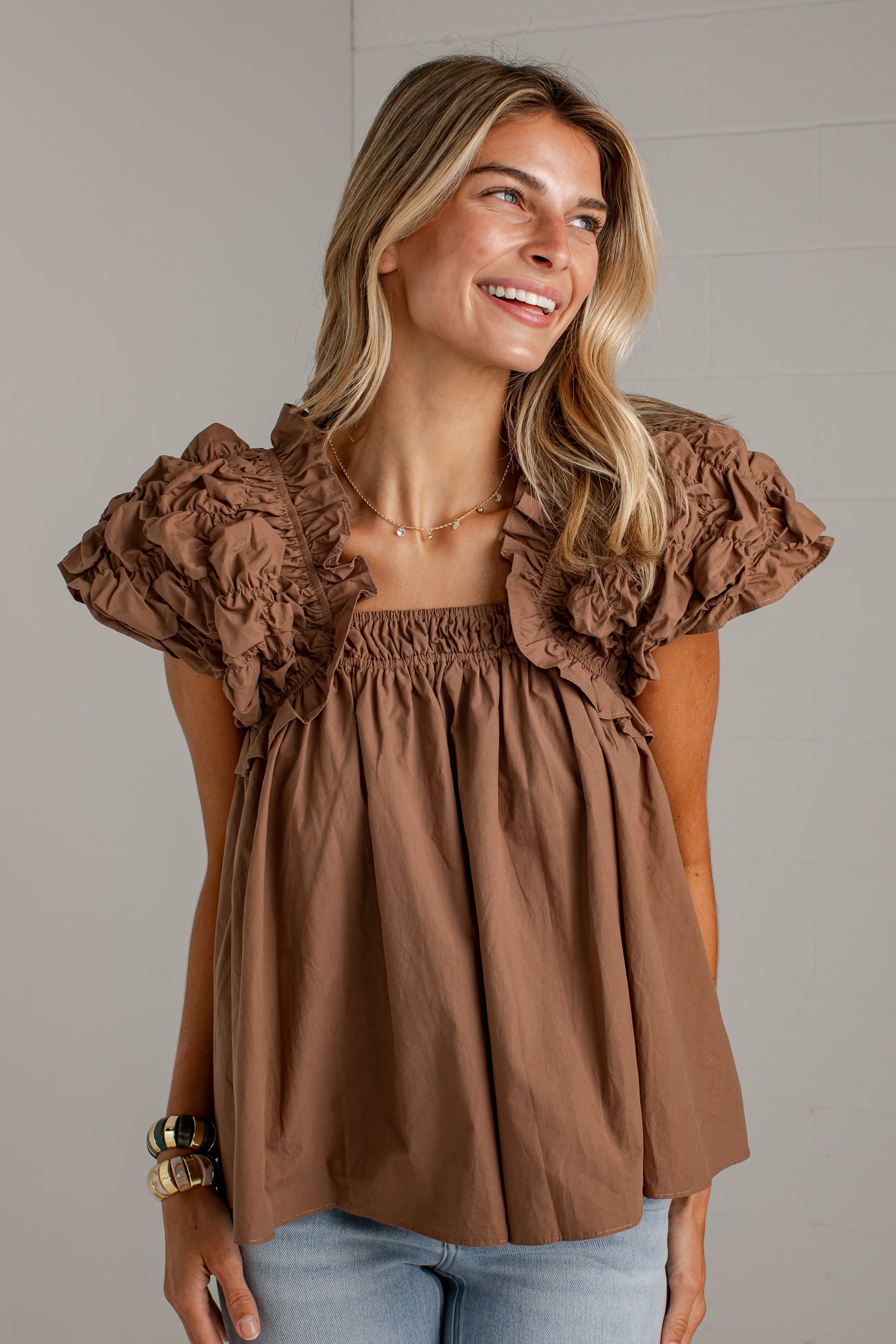 A woman wearing a brown ruffled short-sleeve top with an elasticized neckline and light blue jeans, standing against a light gray paneled wall.