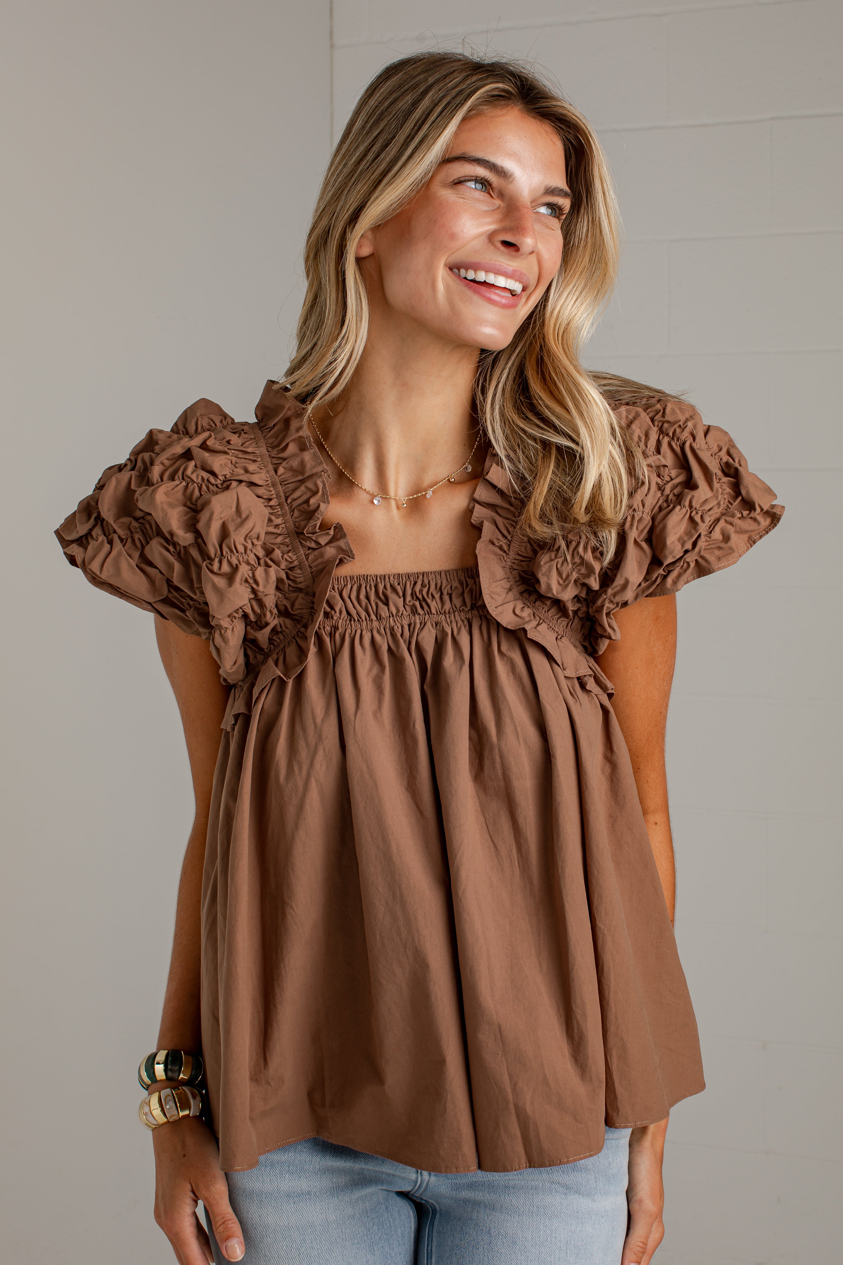 A woman wearing a brown ruffled short-sleeve top with an elasticized neckline and light blue jeans, standing against a light gray paneled wall.