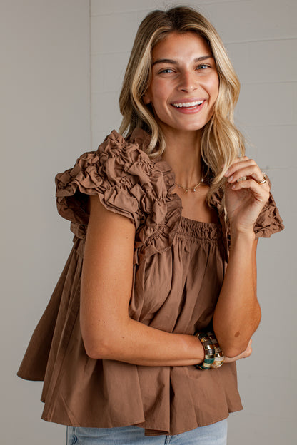 A smiling blonde woman wearing a brown ruffled short-sleeve top and light blue jeans, standing against a light gray paneled wall.