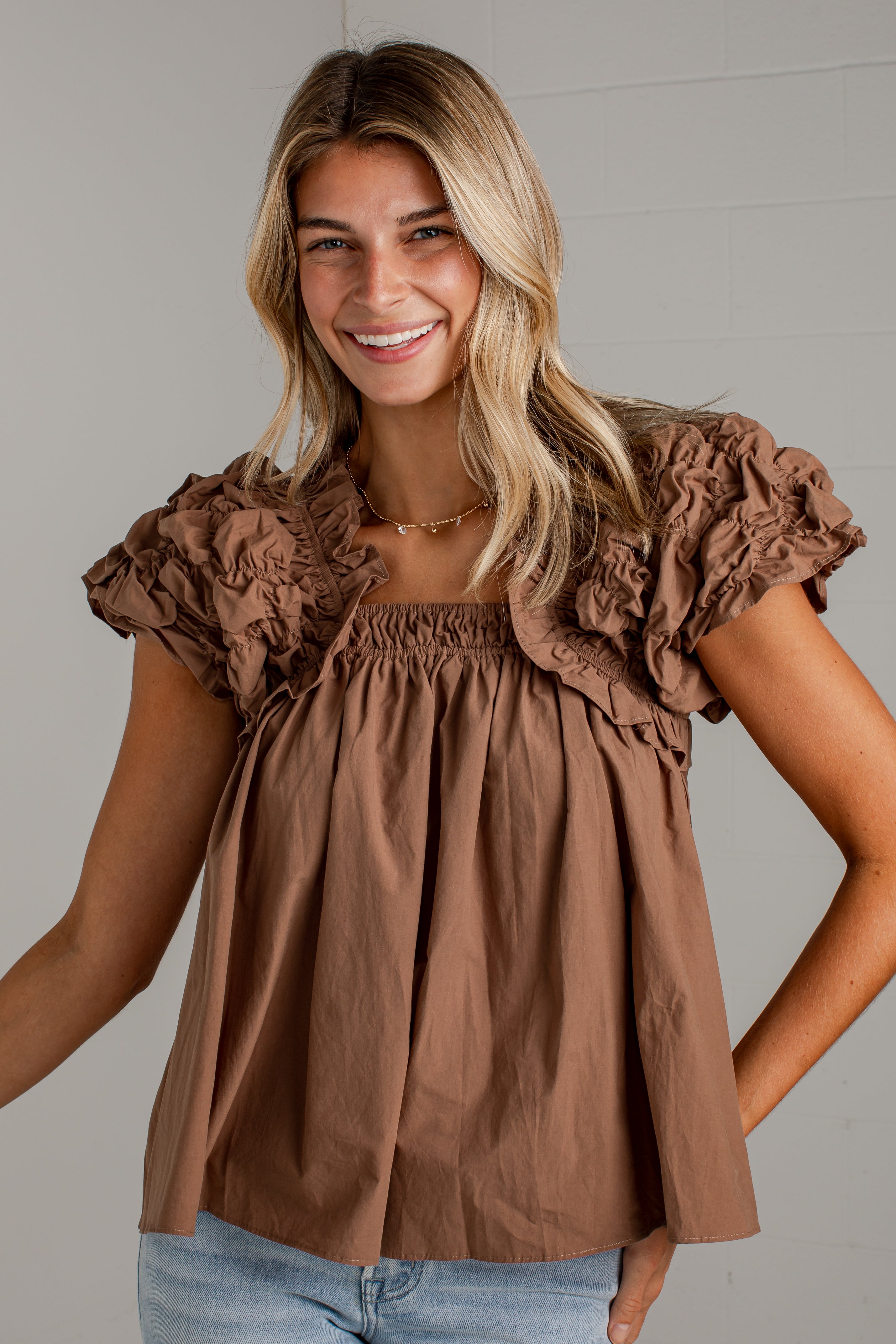 A woman wearing a brown ruffled short-sleeve top with a smocked bodice and puffed sleeves, paired with light blue jeans, standing against a light gray wall.