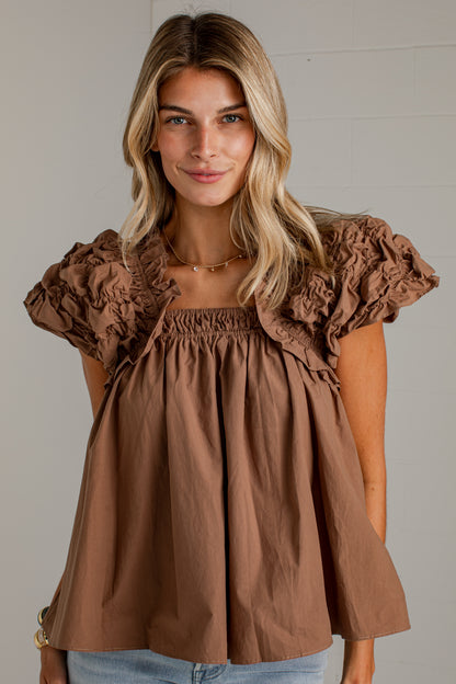 A close-up of a woman wearing a brown smocked-shoulder top with dramatic ruffled puff sleeves and a gathered neckline.
