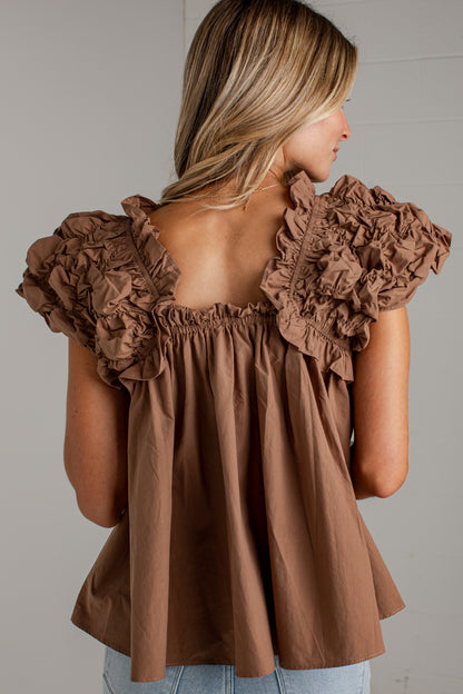 A woman shown from the back wearing a brown ruffled off-the-shoulder top with a smocked neckline and flared peplum-like bottom, paired with light blue jeans against a plain light gray wall.