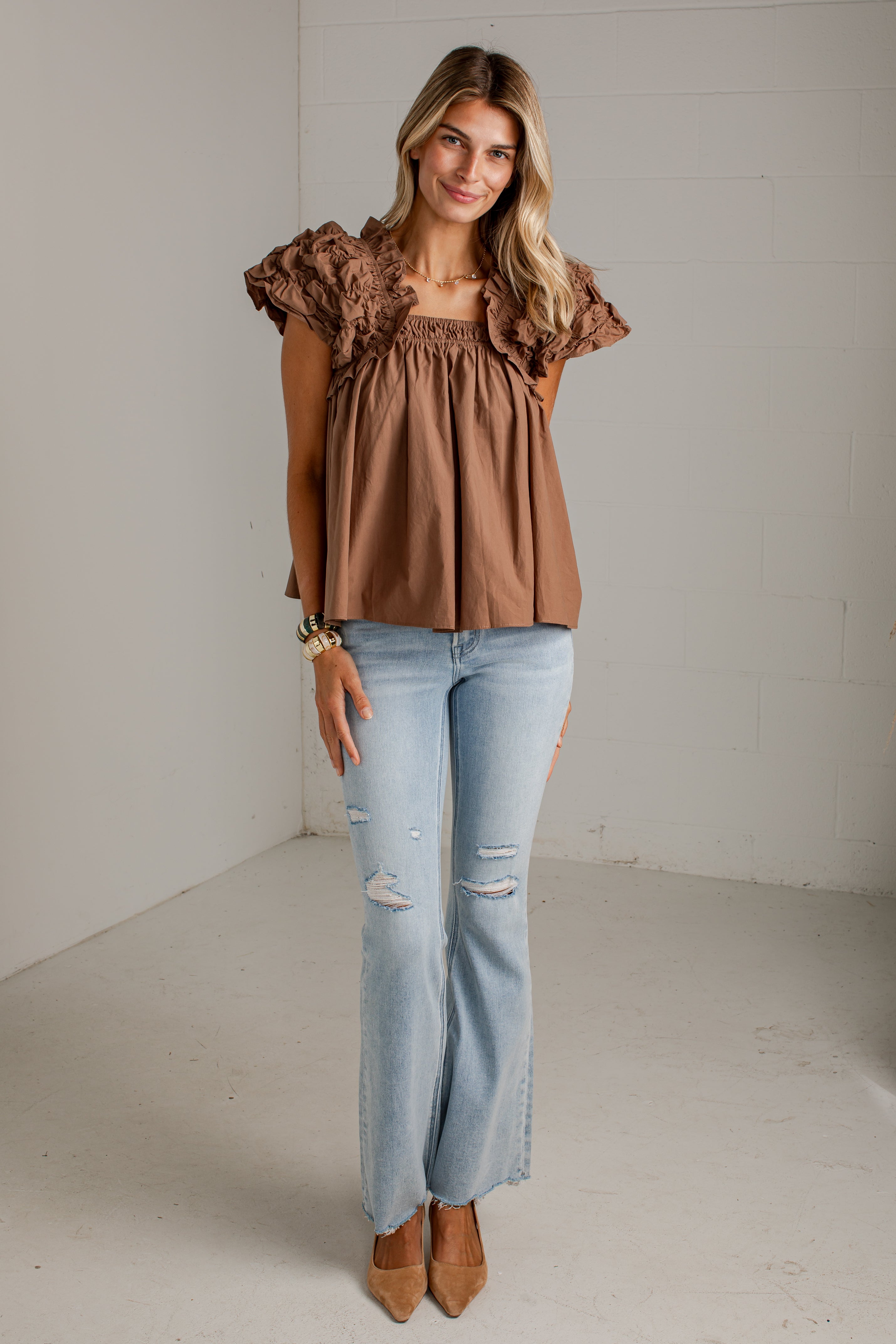 A woman modeling a brown ruffled short-sleeve top with a smocked bodice, paired with light-wash distressed jeans and tan pointed-toe heels, standing in a bright studio with white walls.