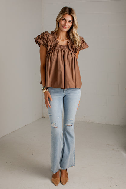 A woman modeling a brown ruffled short-sleeve top with a smocked bodice, paired with light-wash distressed jeans and tan pointed-toe heels, standing in a bright studio with white walls.