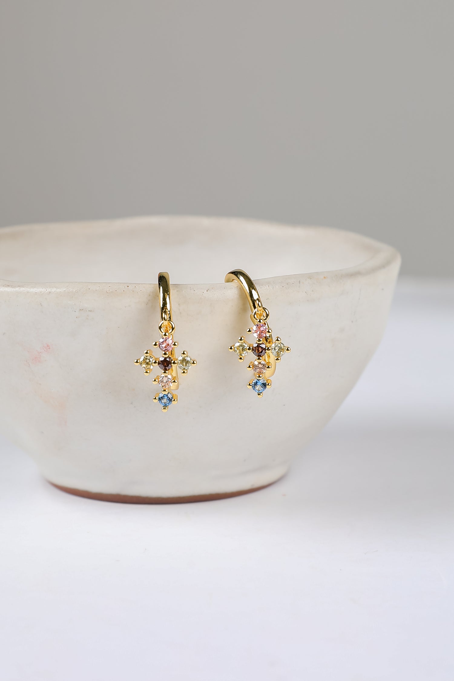 Gold earrings with colorful stones on a white surface with a beige bowl.