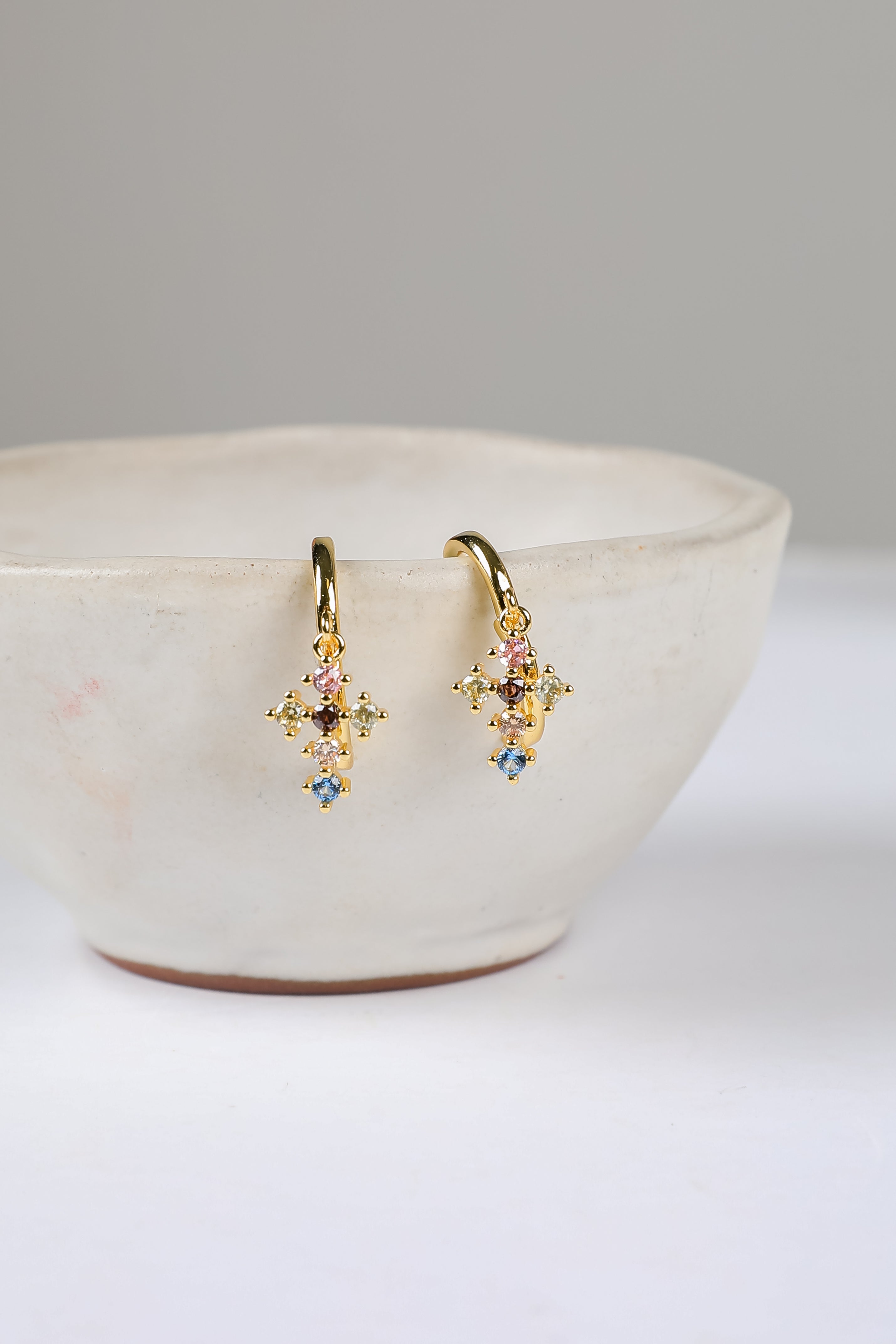 Gold earrings with colorful stones on a white surface with a beige bowl.