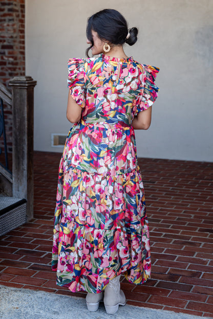 A woman viewed from behind wearing a colorful floral midi dress with ruffled short sleeves and a tiered skirt, standing on a brick patio against a stucco wall.