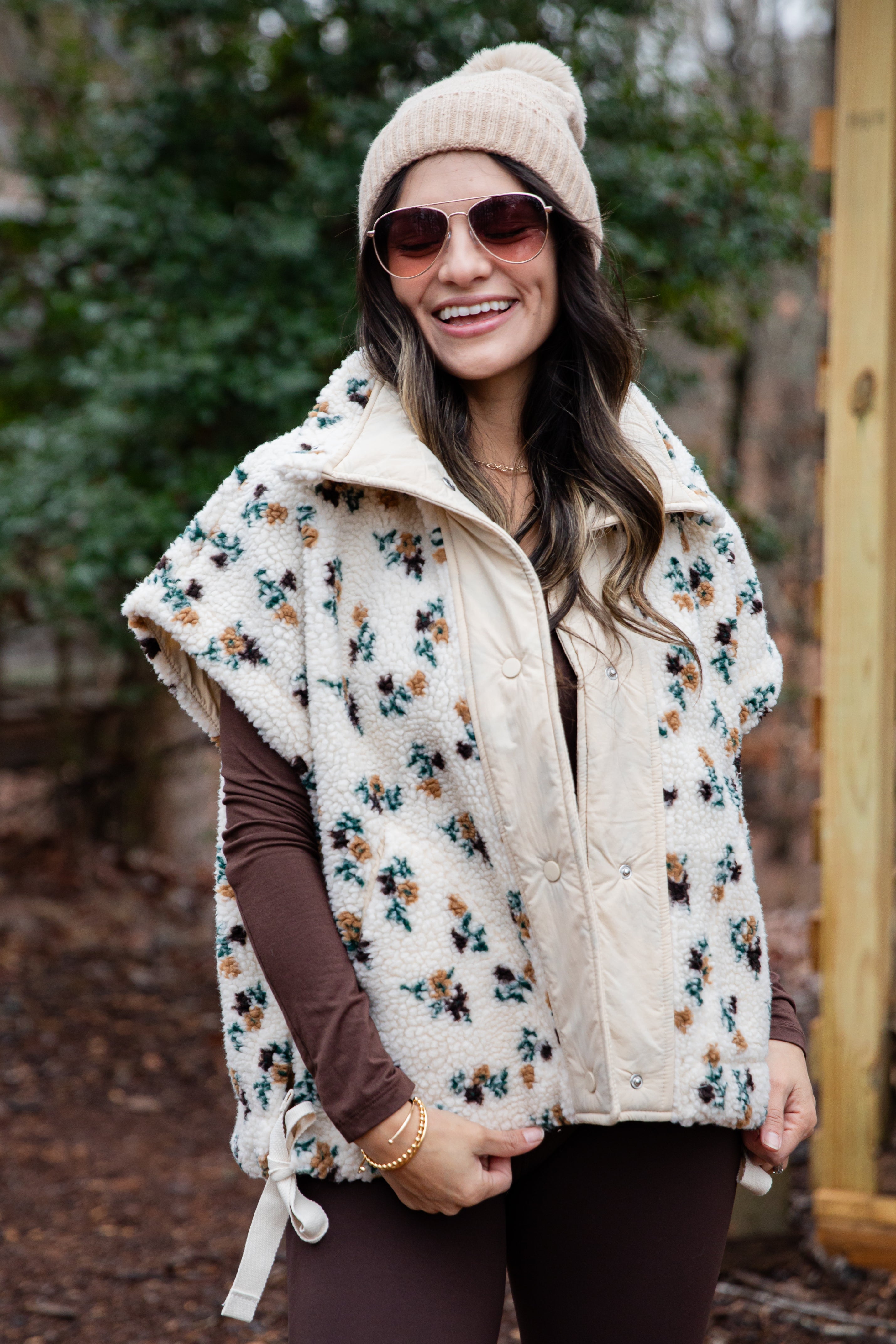 Woman wearing a floral patterned vest over a brown long-sleeve shirt, sunglasses, and a beige knit beanie outdoors.