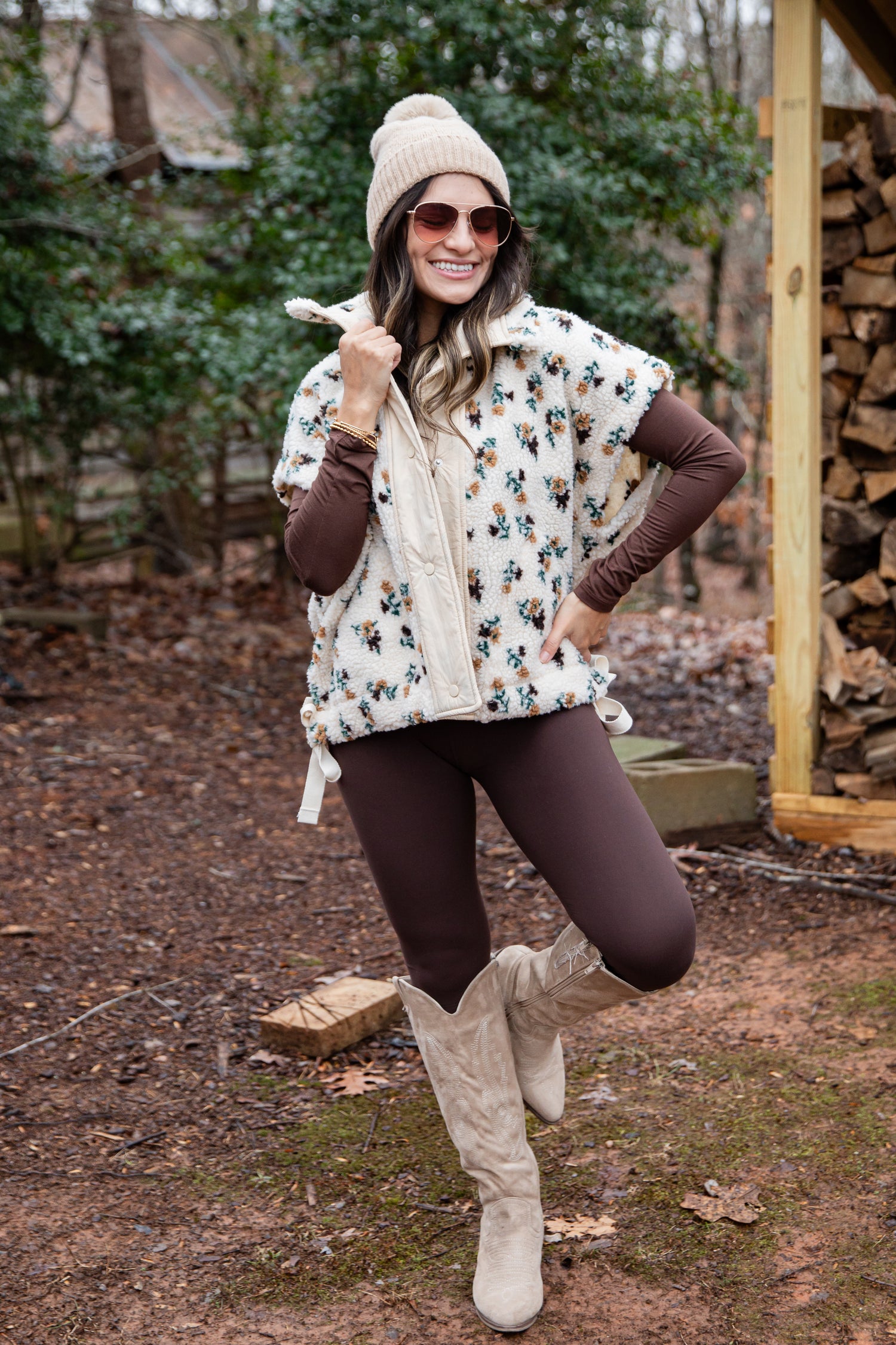 Woman in a floral poncho, brown leggings, and white boots standing outdoors near stacked firewood.