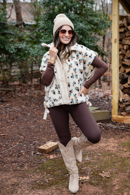 Woman in a floral poncho, brown leggings, and white boots standing outdoors near stacked firewood.
