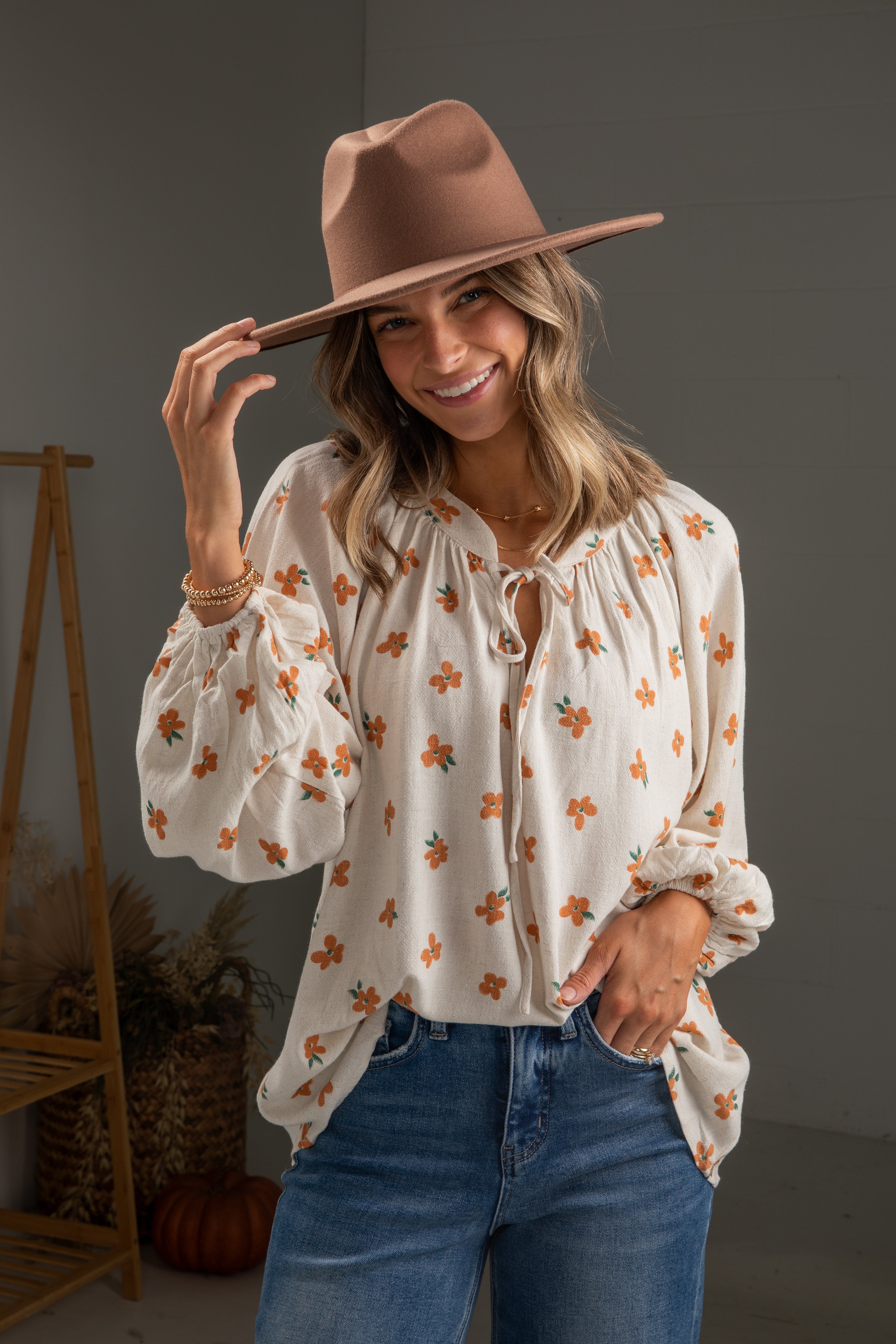 Woman wearing a floral blouse, jeans, and a brown hat indoors.
