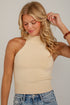 A woman wearing a cream-colored ribbed knit halter crop top and blue jeans, posing against a light gray wall.