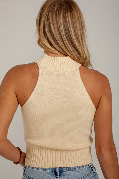 A woman viewed from the back wearing a cream ribbed knit mock neck halter top and blue jeans against a plain light gray background.