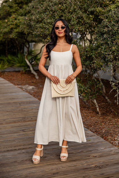 Woman in a white dress holding a beige handbag on a wooden path with greenery.